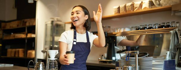Amistosa chica barita sonriente saludando al cliente, sosteniendo su taza de café, orden de comida para llevar preparada, trabajando en la cafetería detrás del mostrador. - Foto, imagen