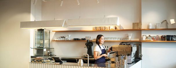 Portrait of cafe counter with barista girl working with coffee machine, making order for client in shop, wearing blue apron. - Photo, Image