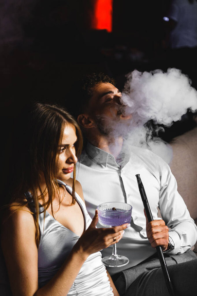Rakastunut pariskunta juo cocktailin ja polttaa vesipiippua terassilla yöllä. Yöelämän käsite - Valokuva, kuva