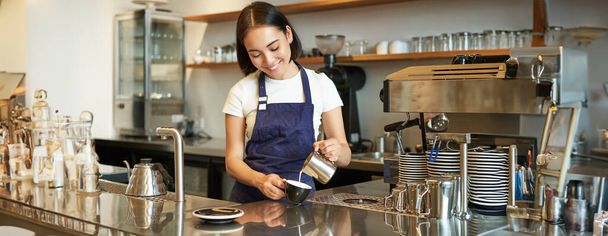 Милая корейская бариста, наливает молоко в кофе, готовит капучино с латте, работает в кафе за прилавком. Люди и рабочее место. - Фото, изображение