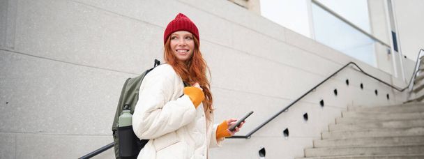 Redhead girl, young woman tourist with backpack, holds smartphone, looks for route on mobile application, searches for hotel on phone map, smiles happily. - Foto, afbeelding