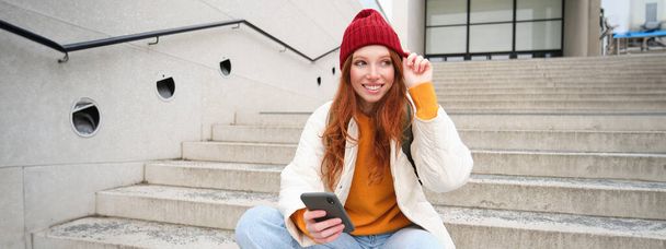 Kaunis punapää tyttö, opiskelija istuu hänen matkapuhelin portaat lähellä kampuksella, hymyilee, pukeutuu punainen hattu, käyttää älypuhelin kaupunki app. - Valokuva, kuva
