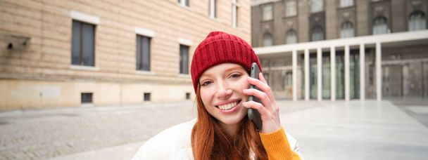 Mobiiliyhteys ja ihmisten käsite. Onnellinen punapää nainen hattu päässä, puhuu matkapuhelimella, soittaa puhelun, käyttää sovellusta soittaa ulkomaille. - Valokuva, kuva