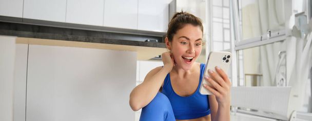 Close up shot of young happy woman, doing sports, workout at home, checking her phone, looking amazed at smartphone screen. - Foto, afbeelding