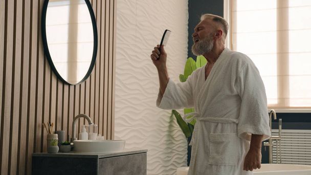 Active happy elderly male in bathrobe at home having fun funny Caucasian old retired senior mature man singing in hair comb after combing hairdo haircare joy pretend sing song pampering with mirror - Photo, Image