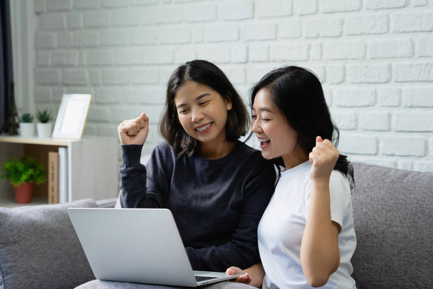 Heureux couple lesbienne asiatique souriant et riant avec de bonnes nouvelles sur ordinateur portable. Ils se sont assis sur le canapé dans le salon. - Photo, image