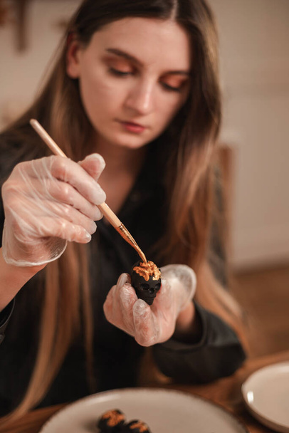Kobieta cukiernik ozdabia cukierki w postaci czarnych czaszek na Halloween ze złota. Proces tworzenia cukierków na Halloween - Zdjęcie, obraz