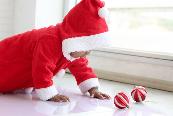 Criança africana no traje vermelho do Papai Noel rastejando para pegar ornamento de bola na sala de estar branca, linda menina celebrando as férias de inverno do Natal. Feliz Natal, infância feliz - Foto, Imagem