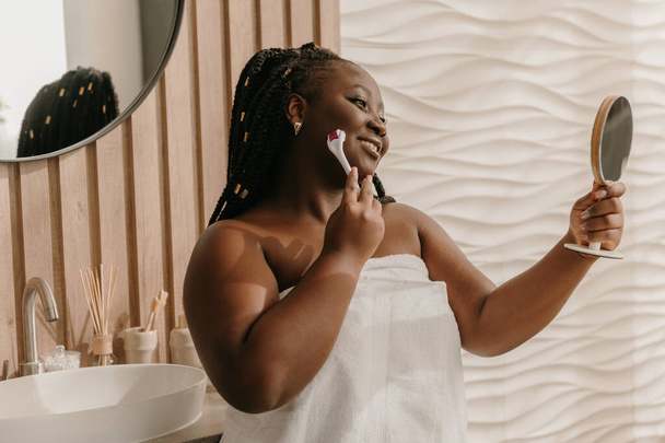 Confiante plus size mulher africana coberta de toalha de massagem rosto com rolo no banheiro doméstico - Foto, Imagem