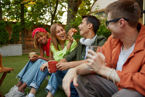 Diverso grupo de jóvenes organizando picnic fiesta al aire libre mientras acampan con furgoneta remolque. Vista de cerca de chicos hipster comer bocadillos dulces y beber té caliente - Foto, Imagen