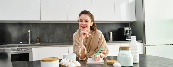 Ελκυστική νεαρή χαρούμενη κοπέλα ψήνοντας στην κουζίνα, φτιάχνοντας ζύμη, κρατώντας βιβλίο συνταγών, έχοντας ιδέες. - Φωτογραφία, εικόνα