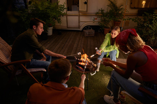 Feliz grupo de amigos asando malvaviscos en la fogata compartiendo historias divertidas por la noche disfrutando de unas vacaciones de glamping al aire libre - Foto, Imagen