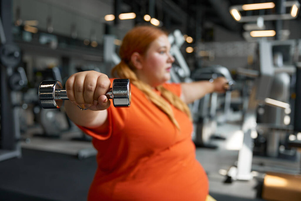 Portrét mladé ženy s nadváhou, která používá činky pumpující svaly na pažích. Obézní dívka cvičení v tělocvičně - Fotografie, Obrázek