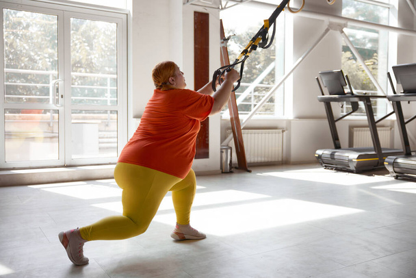 Chubby γυναίκα που εργάζονται σε πρόγραμμα κατάρτισης σωματικού βάρους χρησιμοποιώντας trx βρόχους. Συνολικές ασκήσεις αντίστασης για απώλεια βάρους - Φωτογραφία, εικόνα