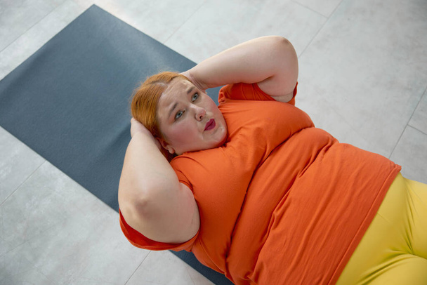 Μια υπέρβαρη γυναίκα που κάνει κοιλιακούς ξαπλωμένη στο χαλάκι. παχύσαρκη κοπέλα που έχει προπόνηση φυσικής κατάστασης στην προσωπική προπόνηση στο γυμναστήριο - Φωτογραφία, εικόνα