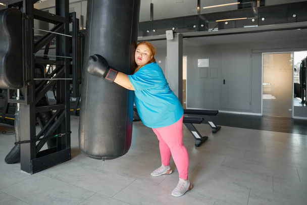 Fatigué femme grosse penché et étreignant sac de boxe tout en s'entraînant dans la salle de gym. Épuisé jeune femme rêvant de chiffre courbé - Photo, image