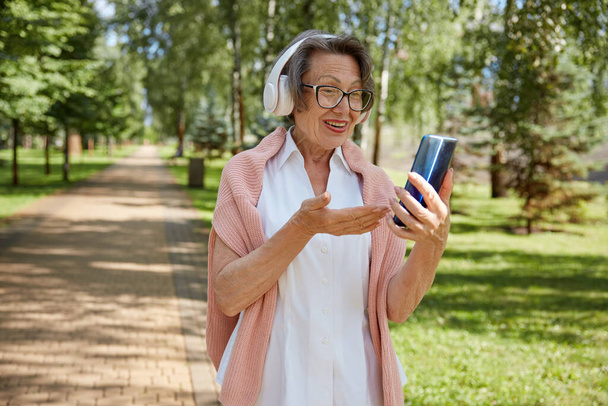Anciana anciana con auriculares que tiene videollamada en línea en un teléfono inteligente utilizando la conexión inalámbrica a Internet con familiares o amigos mientras camina en el parque - Foto, Imagen