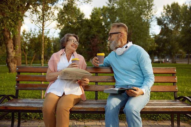 Heureux couple affectueux passer du temps et des loisirs ensemble sur la pension dans le parc. Les conjoints âgés lisent des journaux et des livres, boivent du café et partagent une émotion positive - Photo, image