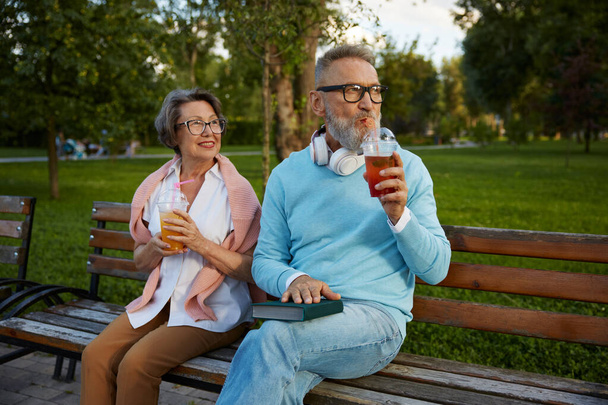Portré nyugodt idős pár iszik frissítő koktélok elvitelre csésze, miközben ül a padon a parkban. Boldog nyugdíjas életet! - Fotó, kép