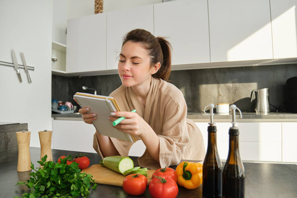 Portret kobiety sprawdzającej notatki kulinarne w notatniku, stojącej w kuchni z warzywami, gotującej jedzenie, przygotowującej pyszną sałatkę z pomidorów, pietruszki i oliwy z oliwek. - Zdjęcie, obraz