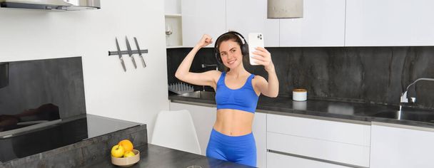 Портрет красивой молодой женщины-блогера, делающей селфи в тренировочной одежде, показывает свои мускулы, сгибает бицепсы на камеру смартфона, записывает видео для социальных сетей. - Фото, изображение