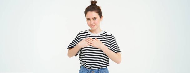 Young smiling woman, feels gratitude, thankful for something, holds hands on heart and looks tender at camera, standing over white background. - Photo, Image