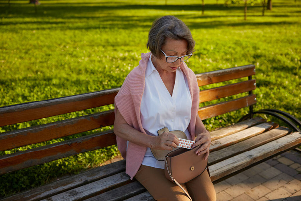 Vecchia donna che prende pillole dalla borsa dopo essersi sentita male a piedi nel parco. Concetto di problema di salute delle persone anziane - Foto, immagini