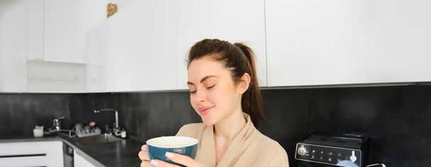 Egy csinos fiatal nő portréja, aki egy csésze kávéval kezdi a napját, a konyhában áll és nagy bögréből issza a cappuccinót, reggelente kedvenc italát fogyasztja.. - Fotó, kép