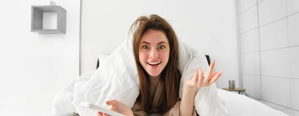 Впечатленная женщина лежит в постели и с помощью мобильного телефона, глядя поражен на камеру контентом онлайн на смартфоне. - Фото, изображение