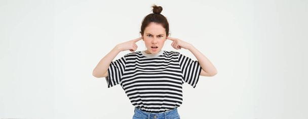 Image of annoyed, bothered woman plugs, shuts her ears with fingers, disturbed by loud noise, stands over white background. - Photo, Image