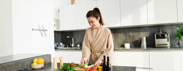 Gesunder Lebensstil. Junge Frau im Bademantel bereitet Essen zu, hackt Gemüse, kocht Abendessen auf Küchentisch, steht vor weißem Hintergrund. - Foto, Bild
