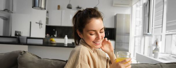 Wunderschöne brünette Frau lächelt kokett, sitzt auf Sofa mit frisch gemachtem Glas Orangensaft, posiert zu Hause im Bademantel, hat ein sanftes Lächeln im Gesicht. - Foto, Bild