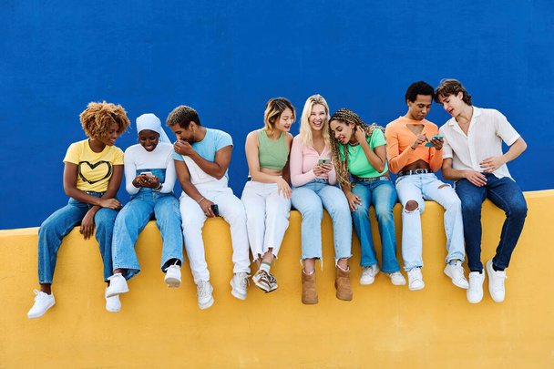 Frontal φωτογραφία των νέων που χρησιμοποιούν το τηλέφωνο κάθεται σε ένα πολύχρωμο τοίχο - Φωτογραφία, εικόνα