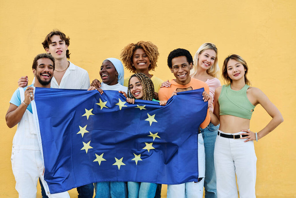 Des amis multiethniques dressent un drapeau de l'Union européenne près d'un mur jaune - Photo, image