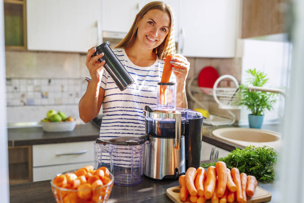 Μια γυναίκα που φτιάχνει χυμό καρότου από φρέσκα καρότα με έναν αποχυμωτή στην κουζίνα. Υγιής τρόπος ζωής - Φωτογραφία, εικόνα