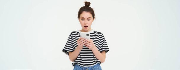 Portret van meisje kijkt geschokt door haar mobiele telefoon, leest een bericht met verbaasd gezicht, geïsoleerd over witte achtergrond. - Foto, afbeelding