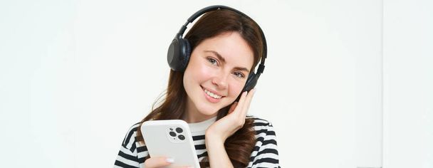 Χαμογελαστή, όμορφη φοιτήτρια με ακουστικά, κρατώντας το κινητό τηλέφωνο, χαμογελώντας και ακούγοντας μουσική, χρησιμοποιώντας smartphone, στέκεται πάνω από το λευκό φόντο. - Φωτογραφία, εικόνα