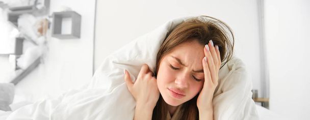 close-up van de vrouw zich onwel voelen thuis, liggend in bed onder witte dekens en fronsen, aanraken van het hoofd, heeft hoofdpijn, migraine, herstellen van covid. - Foto, afbeelding