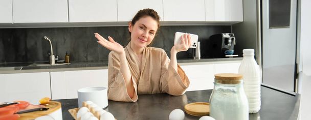 Привлекательная молодая веселая девушка печет на кухне, делает тесто, держит книгу рецептов, есть идеи. - Фото, изображение