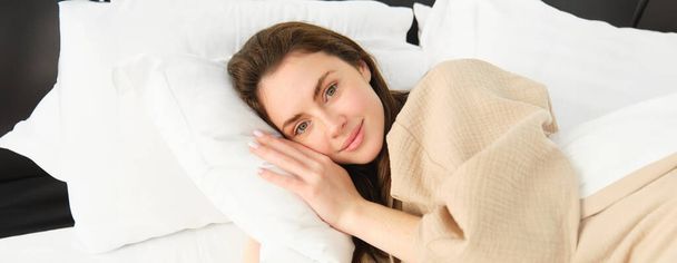 Retrato de una mujer hermosa acostada en la cama, descansando sobre una almohada con los ojos abiertos, mirando a la cámara y sonriendo, usando pijamas, despertando por la mañana. - Foto, Imagen