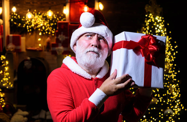 Merry Christmas and Happy New Year. Thoughtful Santa Claus with Christmas present. Pensive bearded man in Santa hat with gift box on Christmas background. Winter holidays celebration. Christmas gifts - Photo, Image