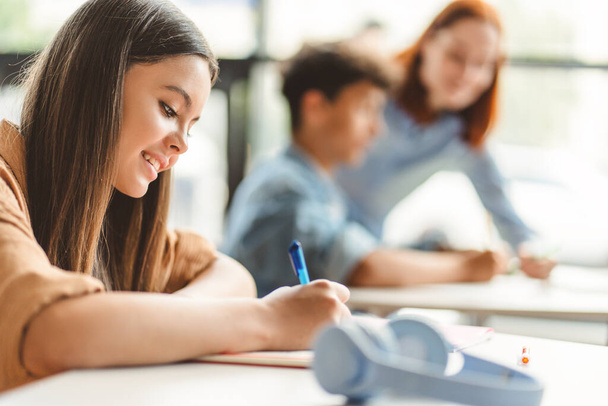 Beautiful, smiling schoolgirl wearing stylish casual clothes sitting at desk, taking notes, holding pen, learning language, exam preparation. Attractive teenage girl in classroom doing homework - Photo, Image