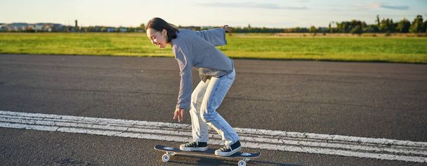 Szczęśliwa dziewczyna skater jazdy na deskorolce i zabawy na pustej ulicy. Uśmiechnięta kobieta korzystająca z przejażdżki krążownikiem po słonecznej drodze. - Zdjęcie, obraz