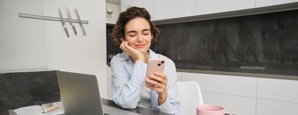 Obrázek mladé ženy, jak doma používá svůj smartphone. Dívka sedí s mobilním telefonem v kuchyni a usmívá. - Fotografie, Obrázek