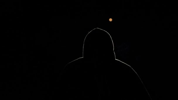 Silhouette einer Person auf schwarzem Hintergrund. Ein Mann mit Kapuze, beleuchtet vom Licht, bläst. Ein Hacker in der Nacht, eine mysteriöse Person, deren Gesicht nicht sichtbar ist. Er raucht eine Zigarette - Filmmaterial, Video