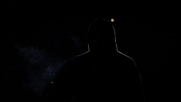 Silhueta de uma pessoa em um fundo preto. Um homem de capuz, iluminado pela luz, sopra. Um hacker à noite, uma pessoa misteriosa cujo rosto não é visível. Ele fuma um cigarro - Filmagem, Vídeo
