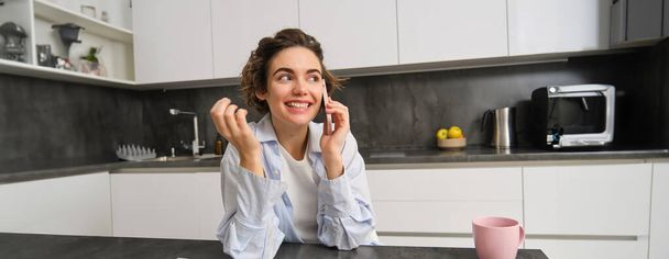 Buněčná technologie a lidé. Usmívající se brunetka sedí doma v kuchyni, mluví na mobilním telefonu. - Fotografie, Obrázek