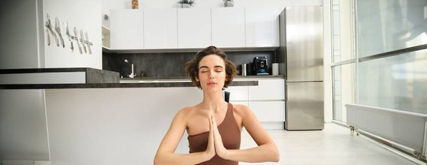 Primer plano retrato de una joven en casa, sentada y meditando, practicar yoga, hacer pose de loto, hacer ejercicio en interiores. Concepto de deporte y bienestar de las mujeres - Foto, Imagen