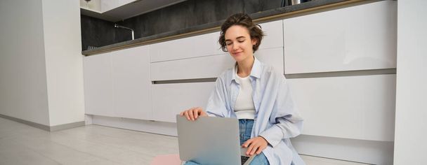 Fiatal nő dolgozik otthonról konyhapadlón, házi feladatot csinál laptopon, internetet használ számítógépen, szabadúszó. - Fotó, kép