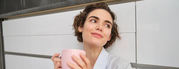 Czułość i relaks. Młoda kobieta pije kawę i uśmiecha się, zrelaksowana filiżanką herbaty, siedzi na podłodze w kuchni. - Zdjęcie, obraz